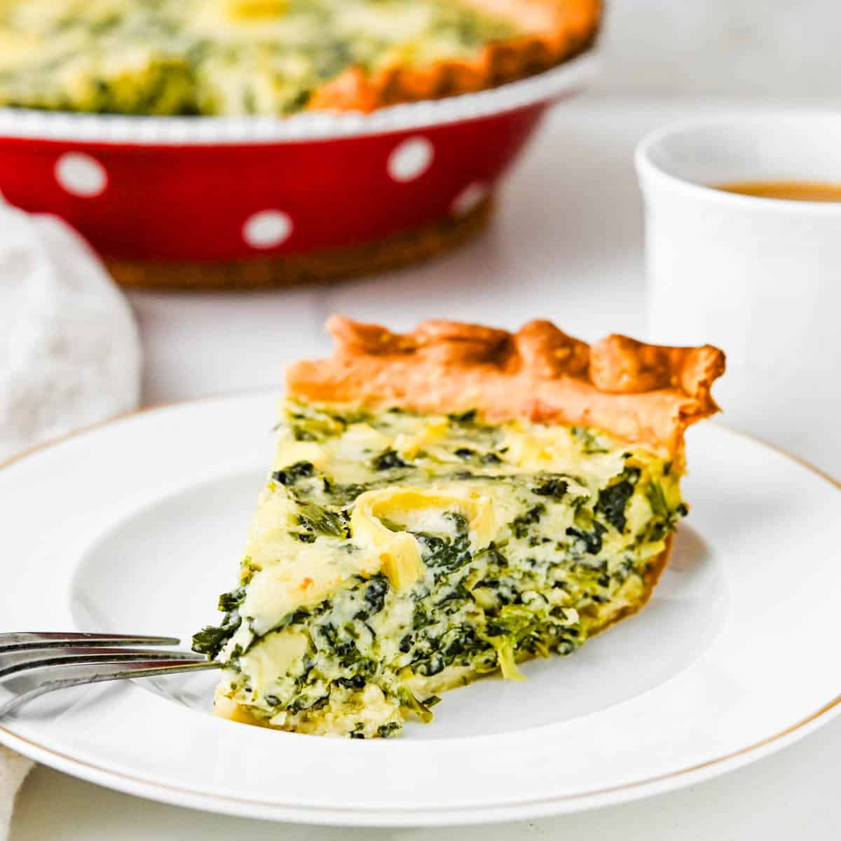 A slice of spinach artichoke quiche on a plate with a cup of coffee.