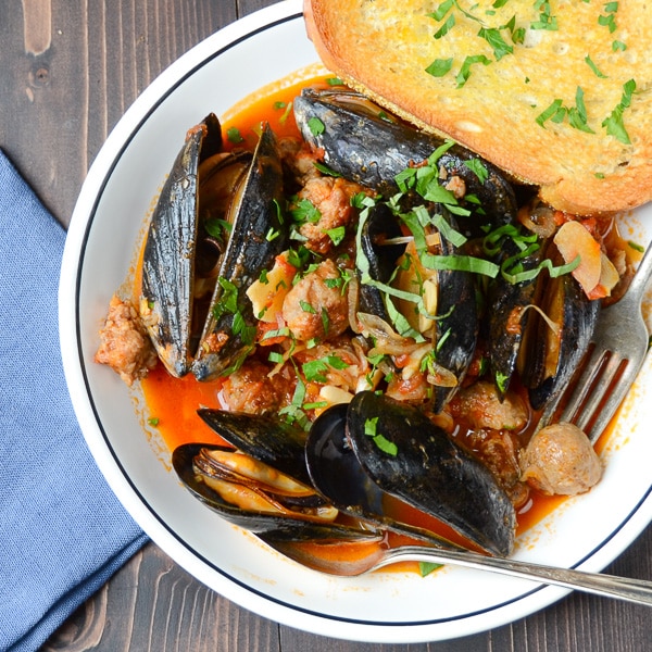 A shallow bowl filled with sausage, mussels and the savory tomato wine broth with a side of garlic bread.