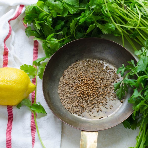 cumin and coriander seed in a skillet with fresh herbs and lemon on the side.