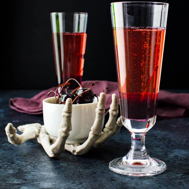 A skeletal hand holding a bowl of amarena cherries next to two bloody bellinis.