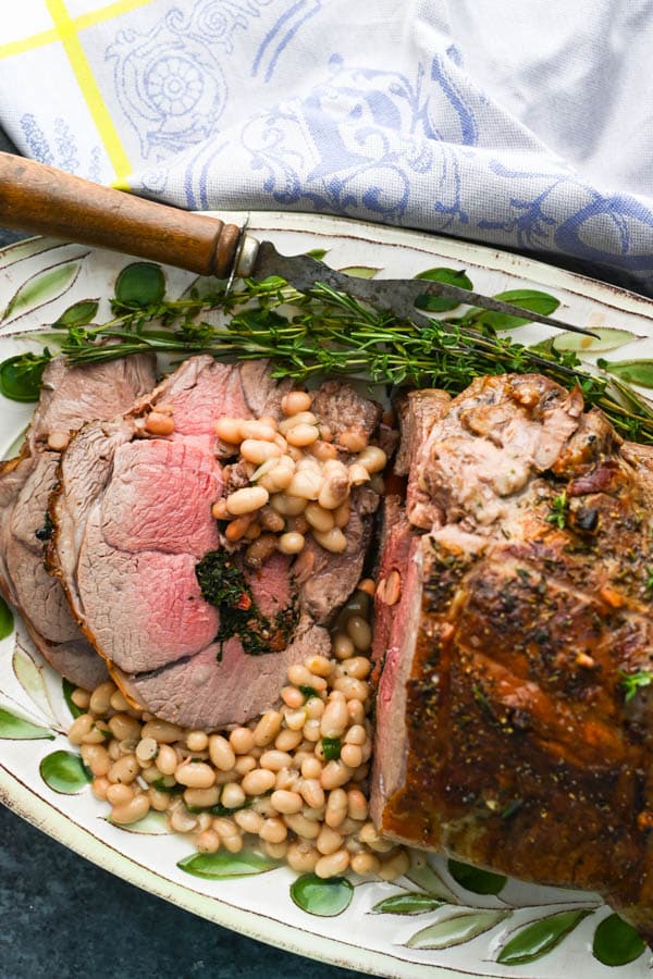 Carved stuffed leg of lamb on a platter with white beans and herbs.