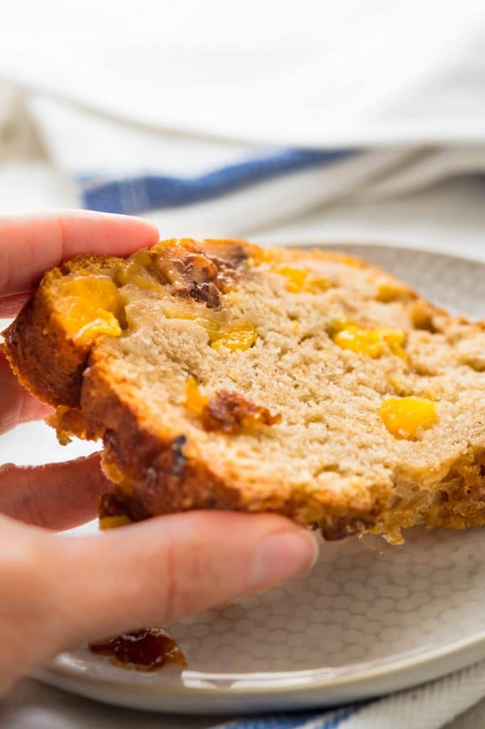 a hand picking up a slice of the peach bread from a plate.