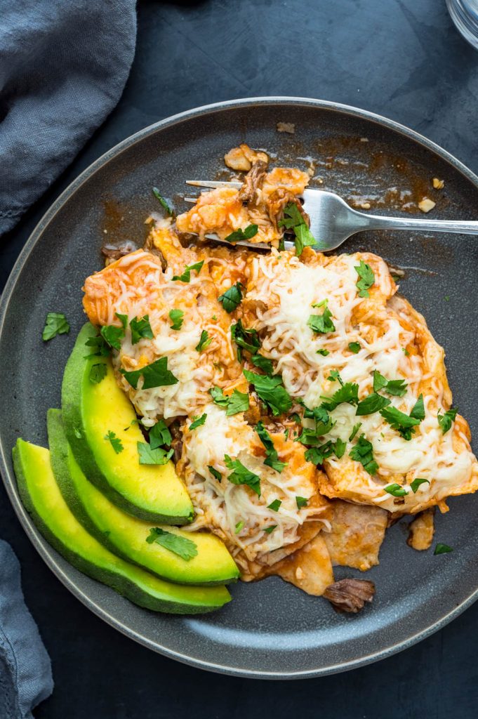 a serving of beef enchiladas on a plate with sliced avocado on the side.