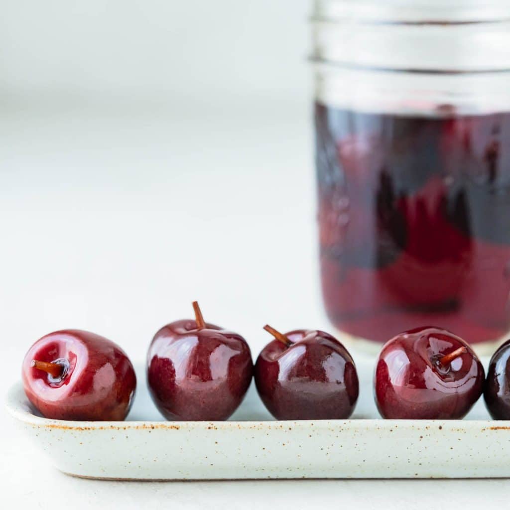 a closeup shot of drunken cherries on a small olive dish with the cherry vodka in a jar behind them.