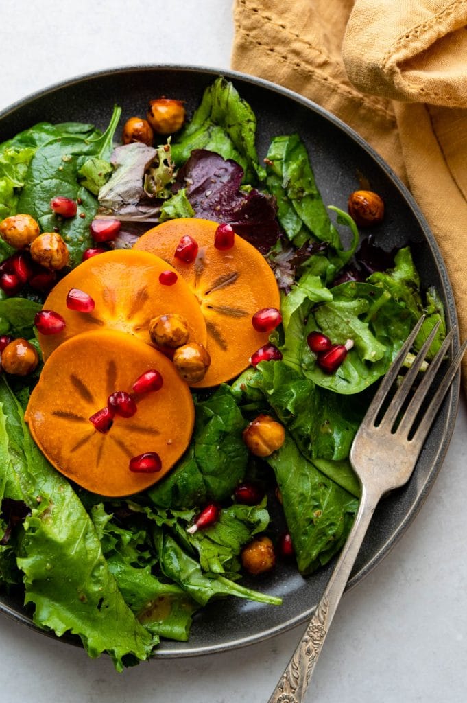 arranging the composed persimmon pomegranate salad on a plate with maple dijon dressing and candied hazelnuts.
