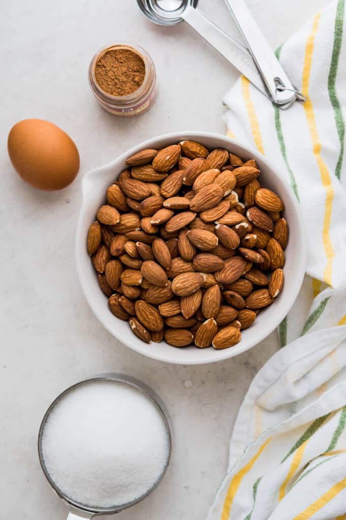 Raw almonds in a bowl with a cup of sugar and egg, cinnamon spice, and a dish towel.