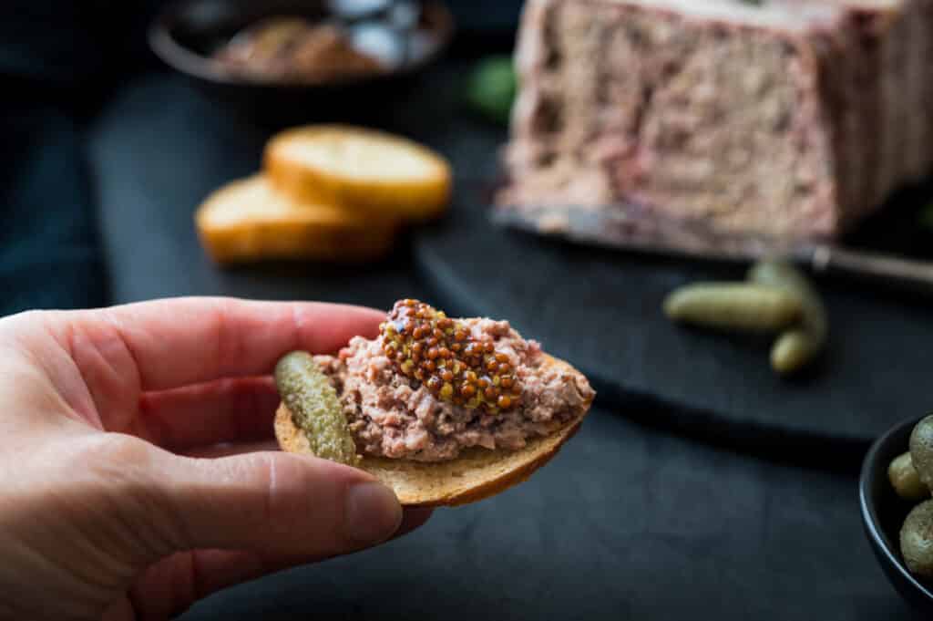 I am holding a cracker with French pate, whole grain mustard, and a slice of cornichon's pickle.