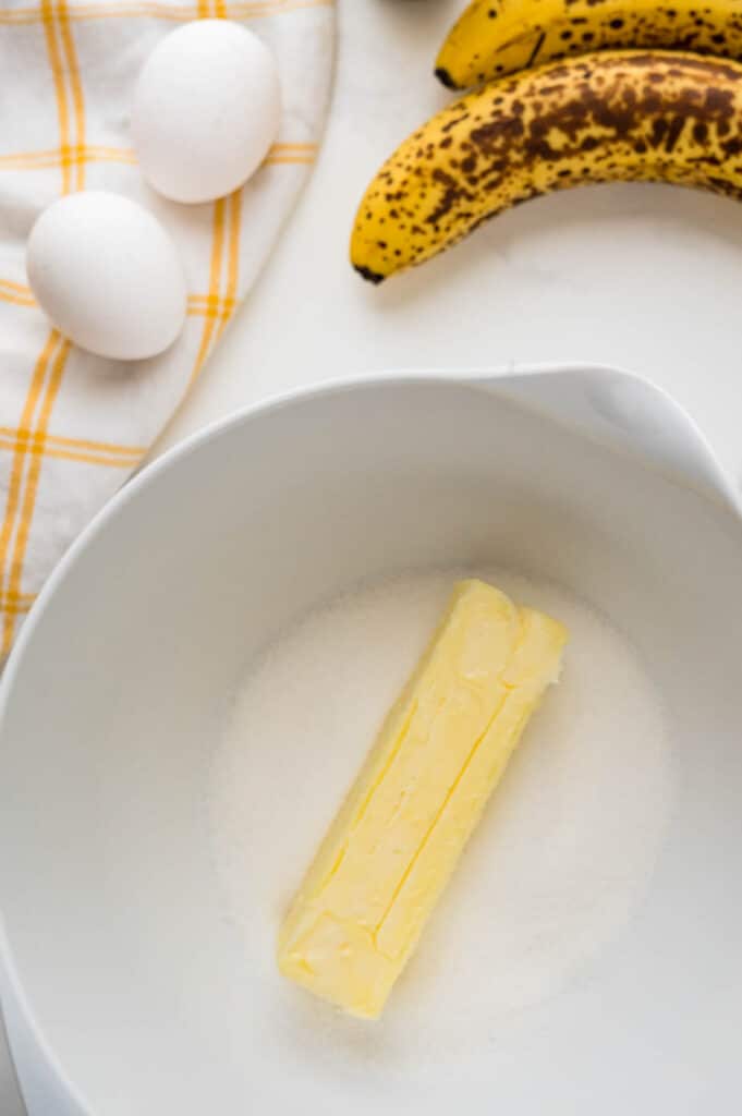 a stick of softened butter in a mixing bowl with granulated sugar. There are two eggs and bananas resting on a kitchen towel to the side.