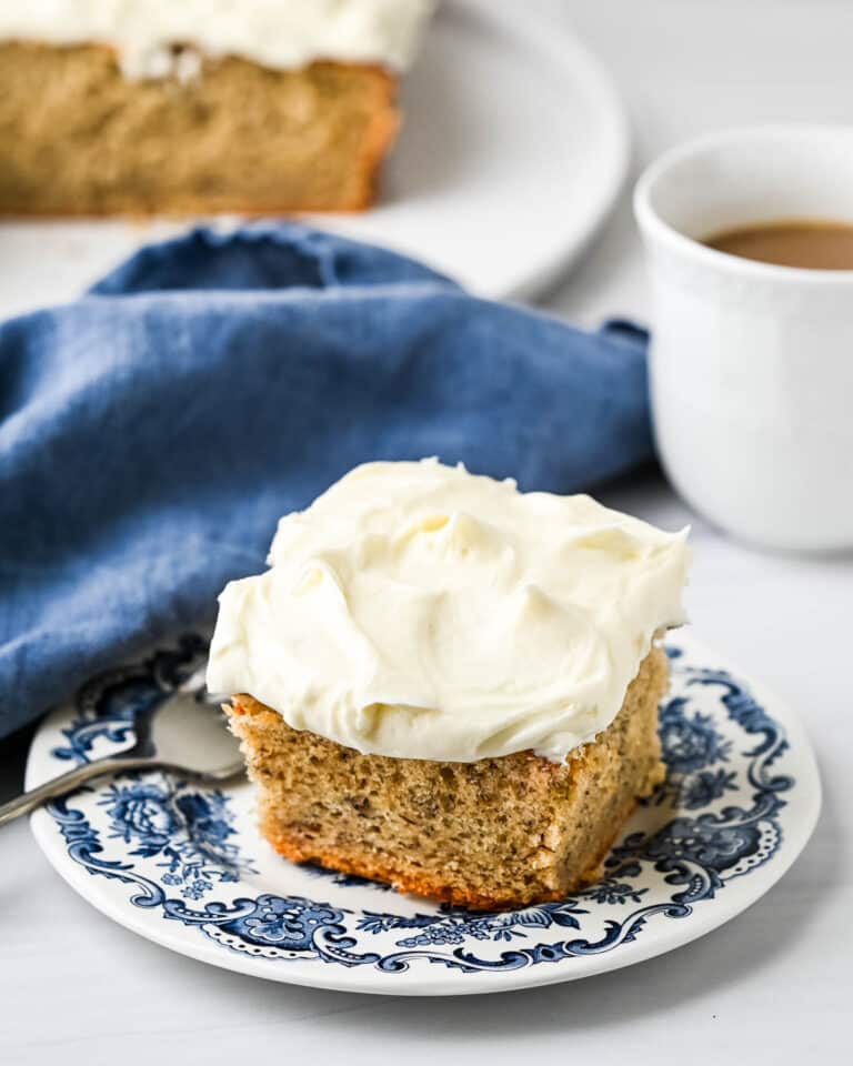 a slice of banana cake topped with cream cheese frosting on a blue plate with a cup of coffee next to it.