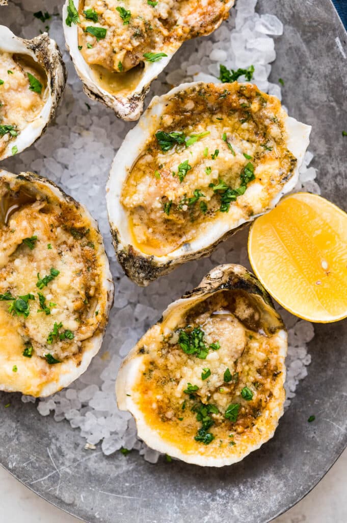 Grilled oysters with cheese and butter topping on a platter lined with rock salt and a wedge of lemon.