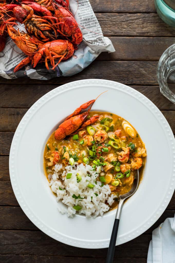 A rustic setting of a white-rimmed bowl filled with the crawfish etouffee recipe with a mound of cooked whole crawfish on a bed of newspapers with a slat-wood background.