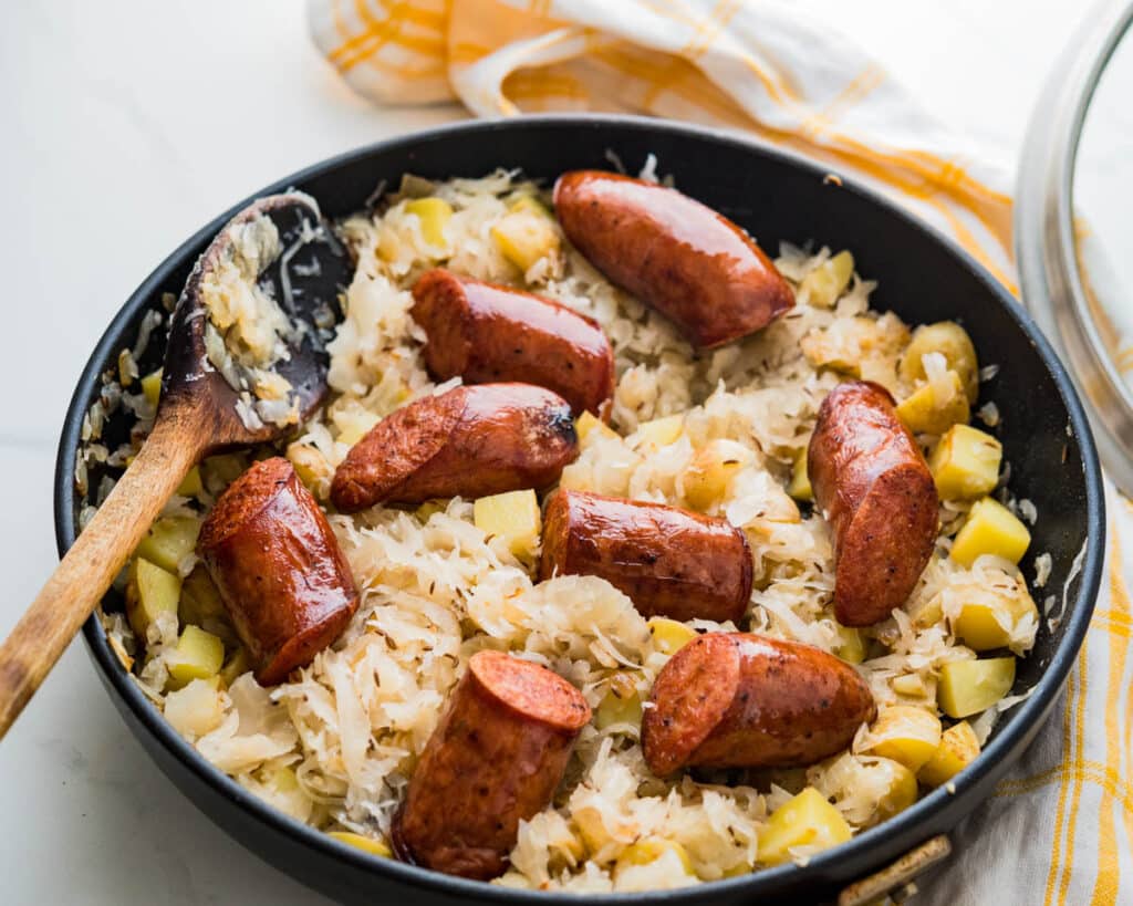 A large skillet filled with simmered sauerkraut and sausage with a wooden spoon to serve.