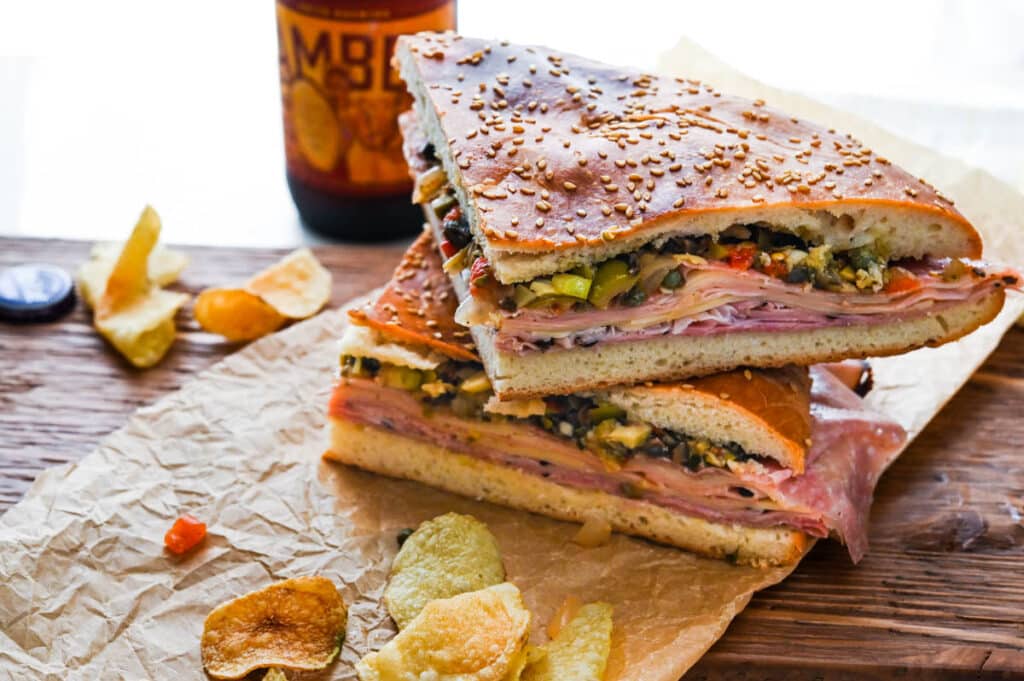 A stack of muffuletta sandwiches with chips and beer on a rustic serving board.