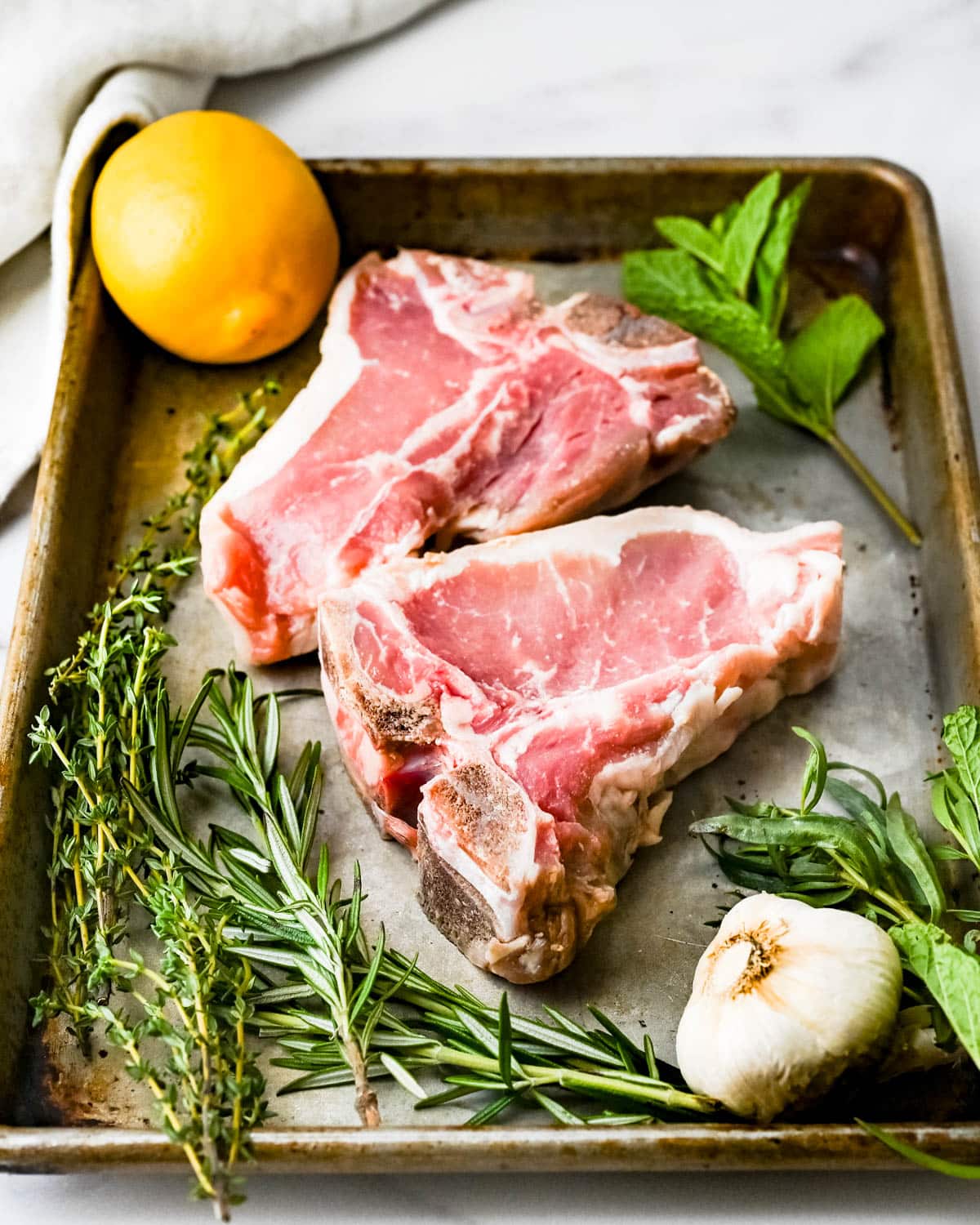 Two t-bone veal chops with fresh herbs on a rimmed baking sheet.