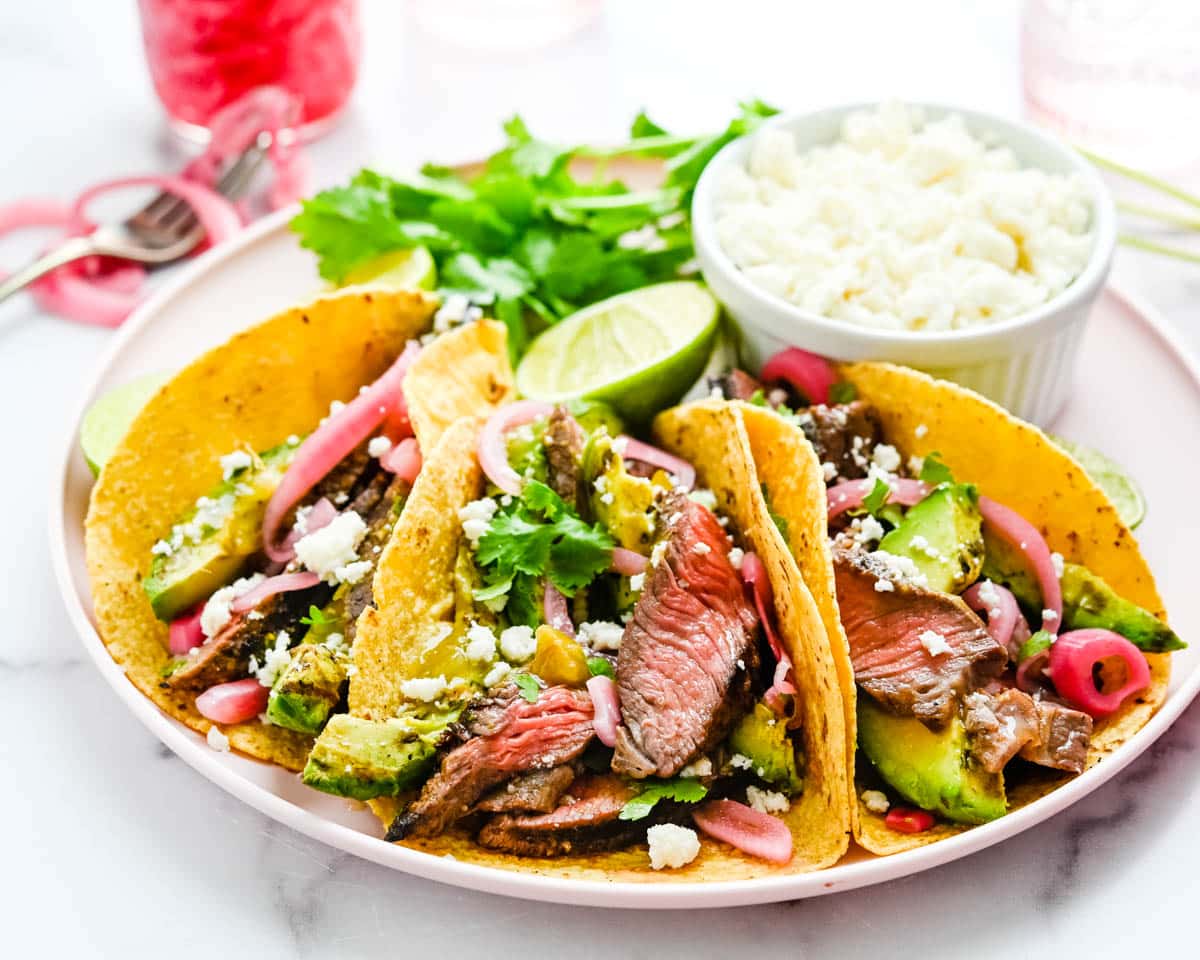 A plate of dressed grilled steak tacos on corn tortillas with grilled avocado and pickled red onion.