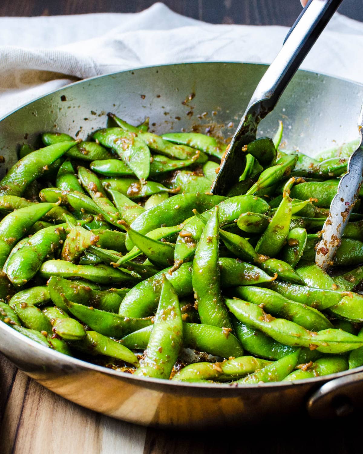 Tossing the edamame with the spicy dressing using a pair of tongs.