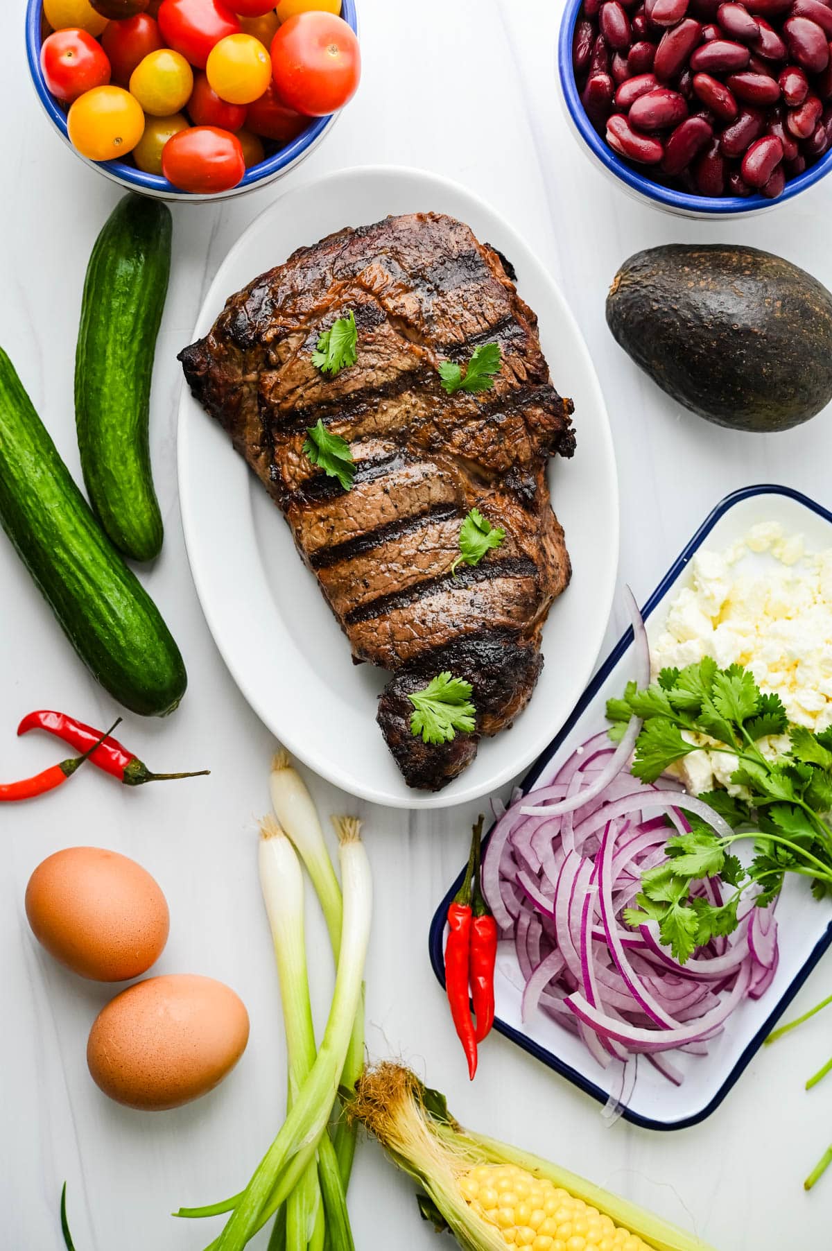 Ingredients for the steak salad include grilled ribeye, tomatoes, kidney beans, cucumber, avocado cilantro, queso fresco, green onions, chilies and jammy eggs.