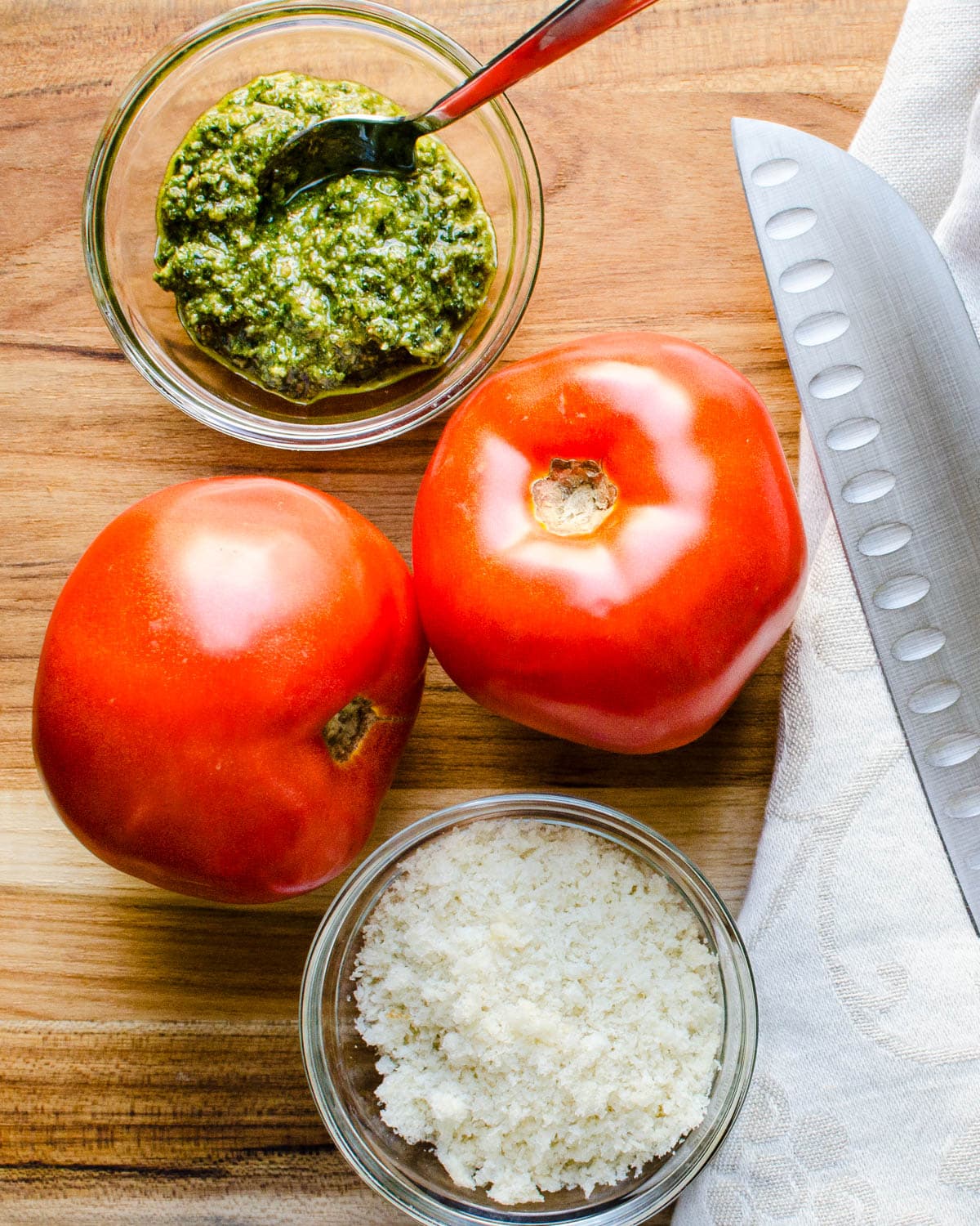 Large beefsteak tomatoes on a cutting board with pesto sauce and breadcrumbs.
