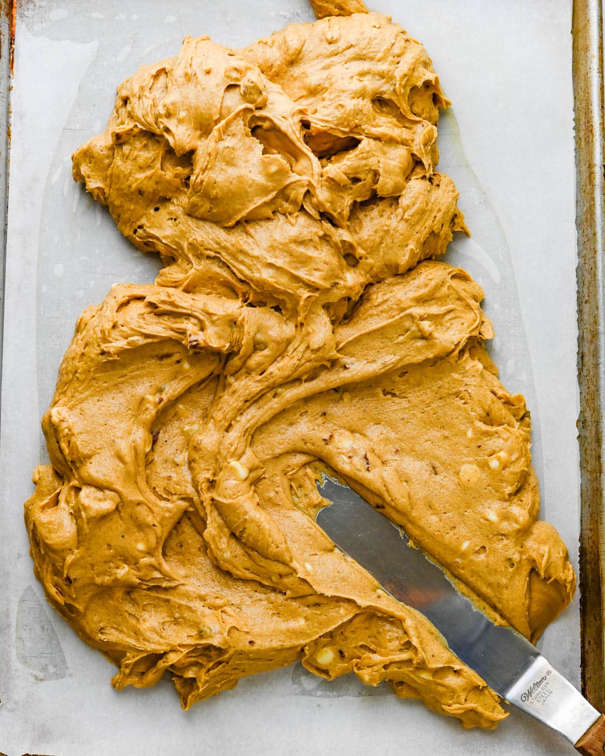 Using an offset spatula to spread the thick pumpkin blondie batter evenly in a prepared half sheet pan.