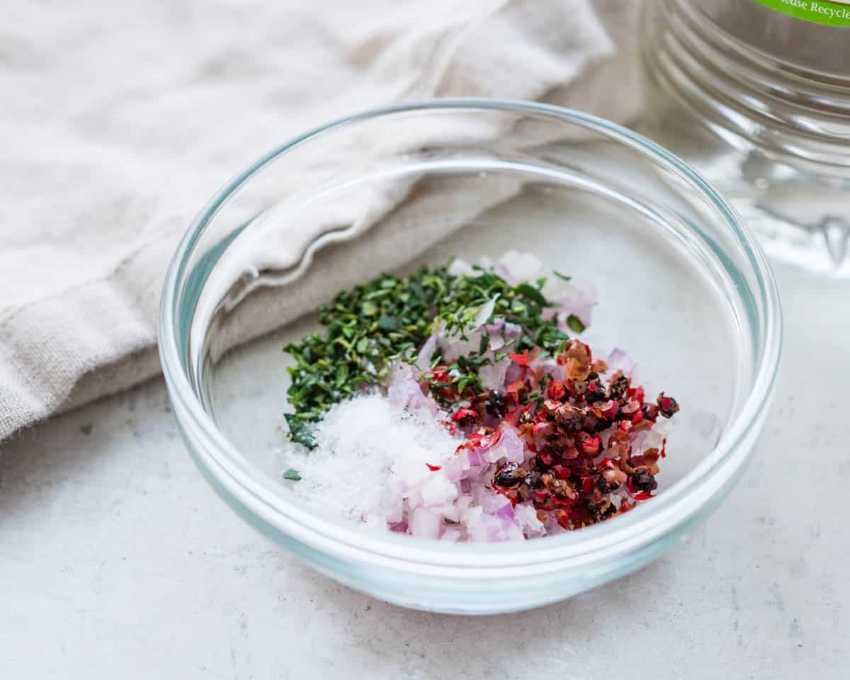 A glass bowl with shallots tarragon, peppercorns and salt.