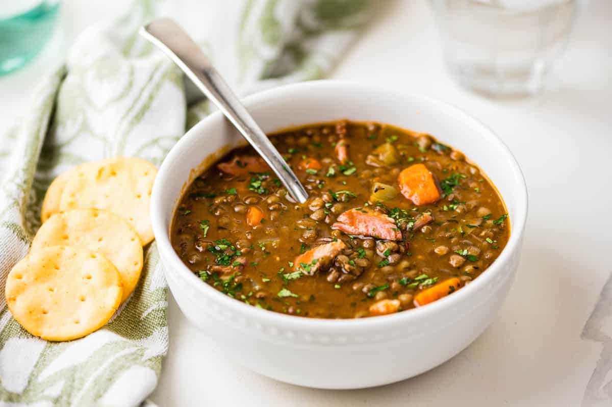 A bowl of ham and lentil soup with crackers on the side.