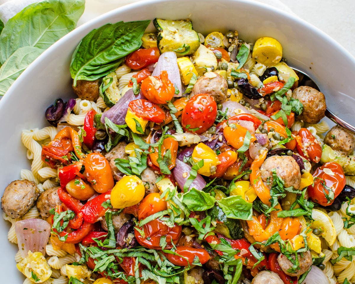 A white bowl with colorful sausage-and-pepper pasta and fresh herbs.