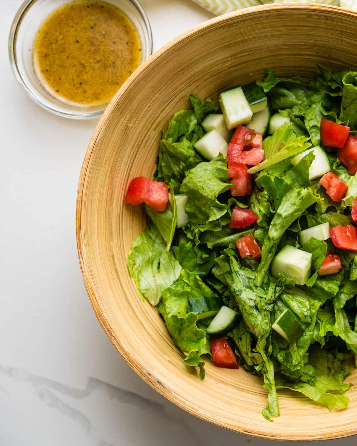 A bowl of green lettuce salad with cucumber and tomato and a bowl of red wine vinaigrette next to it.