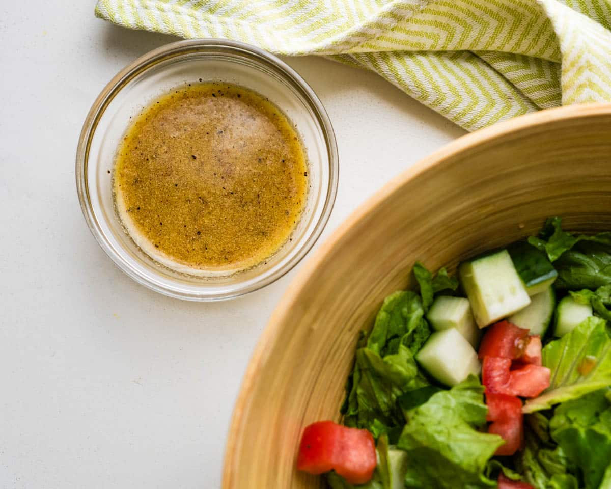 A green salad with lettuce, tomatoes and cucumbers with a bowl of the red wine vinaigrette next to it.